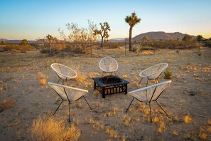 A desert landscape with three white wicker chairs and a small table, surrounded by dry grass and sparse vegetation.
