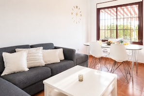 A modern living room with a grey sofa, white cushions, a white coffee table, and a dining area with a table and chairs.