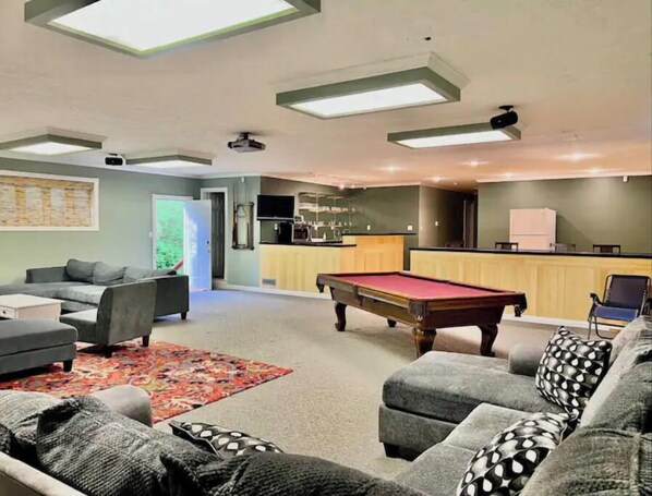 A modern living room with a pool table, grey sofas, and a kitchen area.