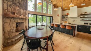 Un comedor amplio con una gran mesa de madera y sillas, una chimenea de piedra y vistas al exterior a través de grandes ventanales.