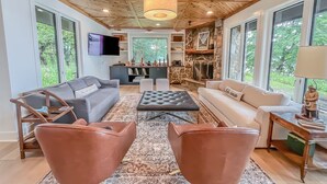 Un salón amplio con techo de madera, ventanas grandes, chimenea de piedra y muebles modernos.