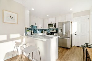 Uma cozinha moderna com armários brancos, uma ilha central com banquetas, uma geladeira de aço inoxidável e um micro-ondas.