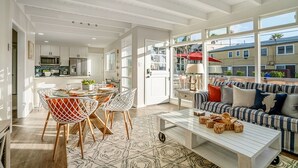 A spacious living area with a dining table, chairs, a sofa, and a coffee table. There is a kitchen in the background with cabinets and appliances. - "WAVE POOL" HOUSE! 1-Short Block to Ocean! (Oceanside)