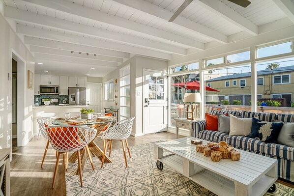A spacious living area with a dining table, chairs, a sofa, and a coffee table. There is a kitchen in the background with cabinets and appliances.
