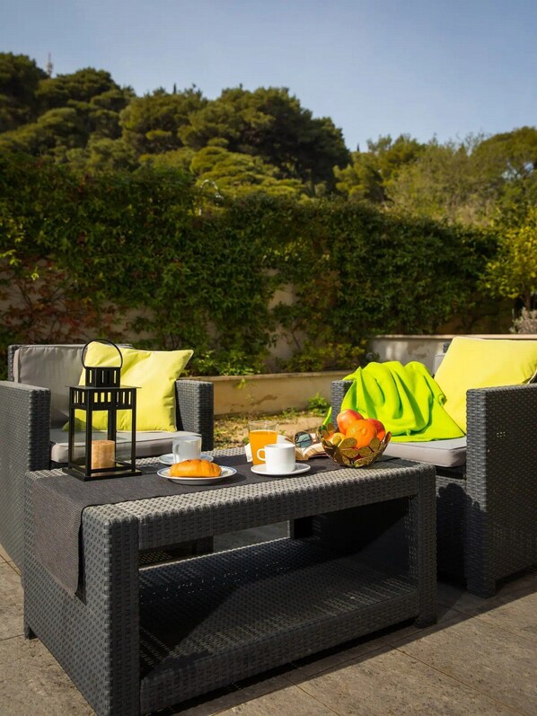 A wicker outdoor seating area with a table set for breakfast, including a croissant, orange juice, and a fruit bowl.