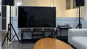 Una sala de estar moderna con un televisor de pantalla plana grande sobre un soporte de madera, dos parlantes en pedestales, una mesa de centro redonda de madera y un sofá gris.