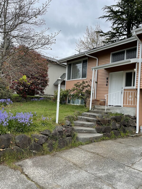 A two-story house with a front porch, a small garden with purple flowers, and a mailbox.