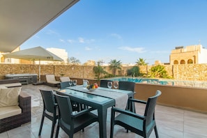 An outdoor patio with a dining table, chairs, a grill, and a swimming pool in the background.