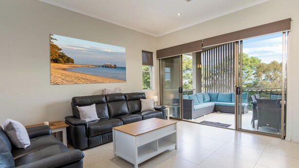 A modern living room with a black leather sofa, a wooden coffee table, and a large sliding glass door leading to an outdoor seating area. - Awesome, modern, open-plan 3rd floor (with lift) unit with stunning water views. (Bongaree)