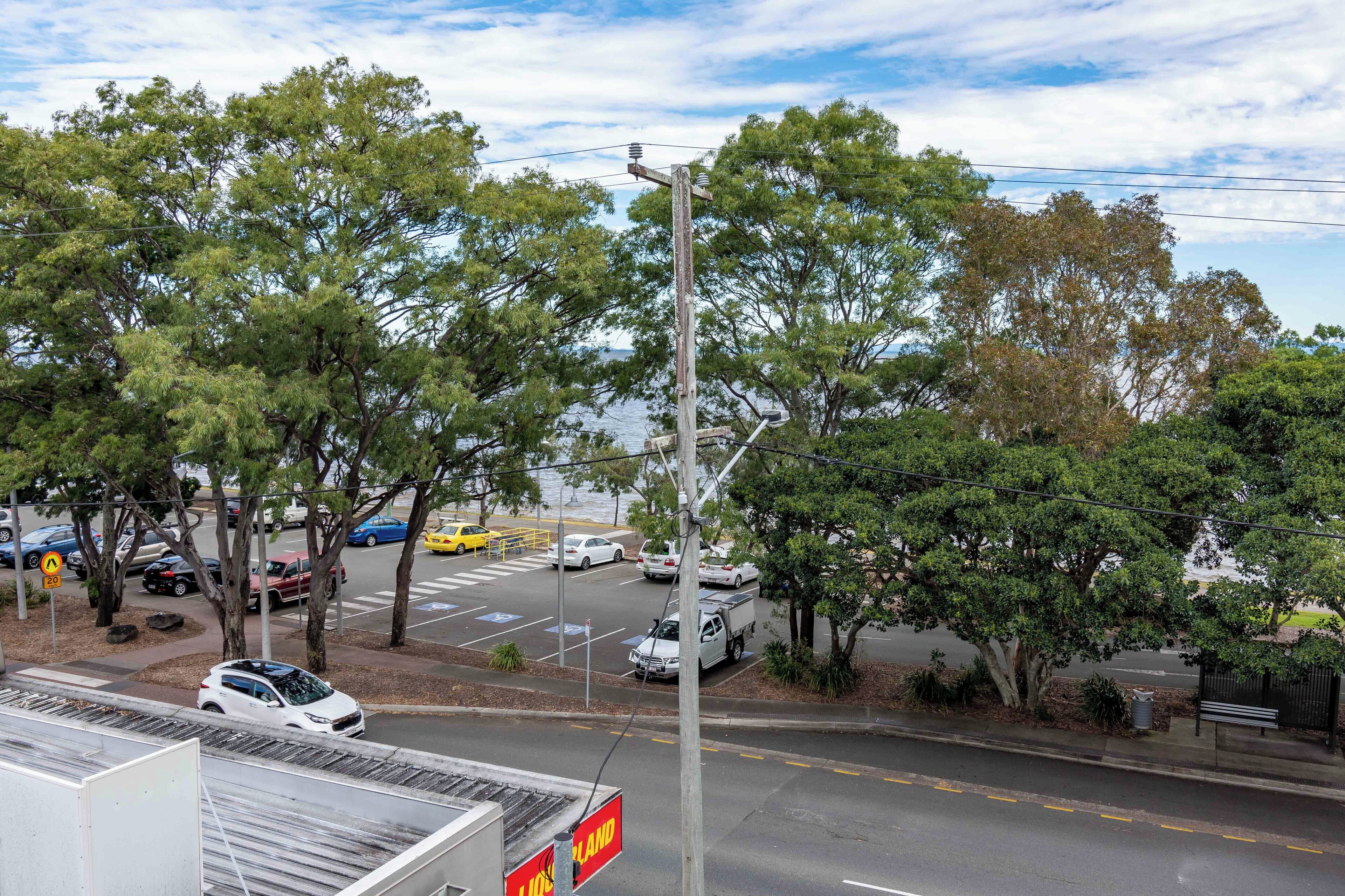 Um estacionamento com carros e um caminhão, rodeado por árvores e um corpo de água ao fundo.