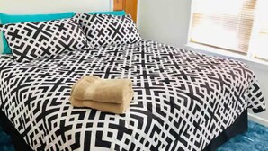 A neatly made bed with a geometric patterned bedspread, a folded towel, and a window with blinds. - Location!! Close to Everything! Spacious and Fun! (Lexington)