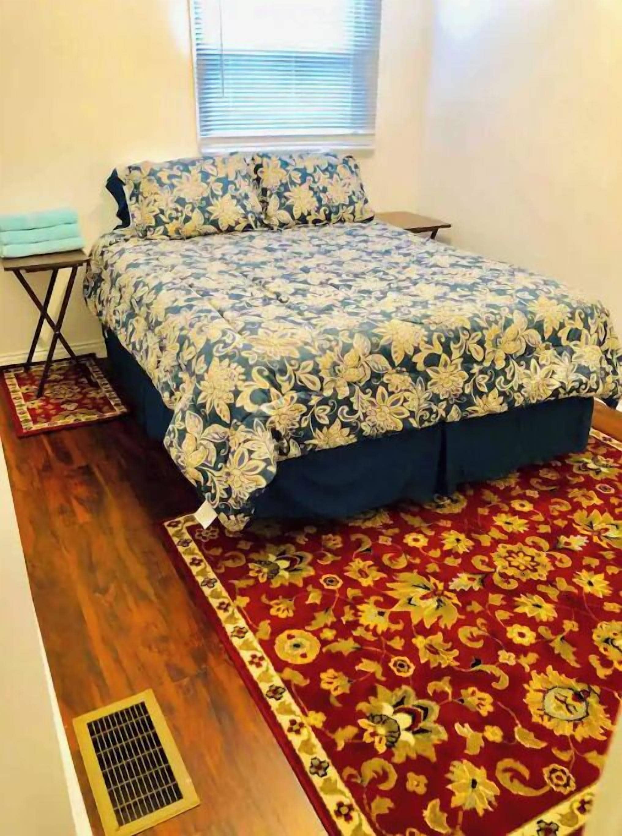 A bedroom with a floral bedspread, a red and gold patterned rug, and a window with blinds.