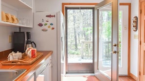 Een keuken met een koffiezetapparaat, mokken en een koelkast versierd met decoratieve magneetjes. Een deur leidt naar een terras met uitzicht op bomen.