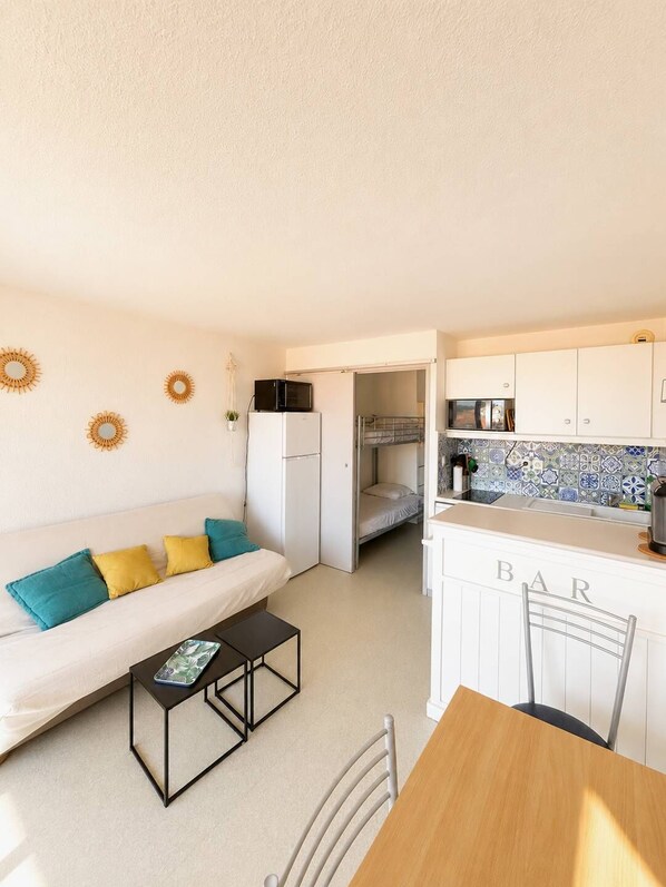 Un espace de vie compact comprenant un coin cuisine, un canapé avec des coussins, une table à manger et une petite table d’appoint.