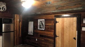Interior d'una cabana rústica amb parets de fusta, un nevera, una porta i una làmpada al sostre.