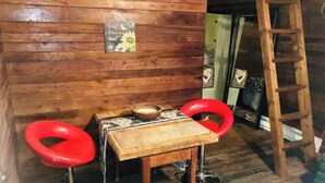 Interior d'una cabana de fusta amb una taula, dos tamborets vermells, una escala i un objecte decoratiu petit a la paret.