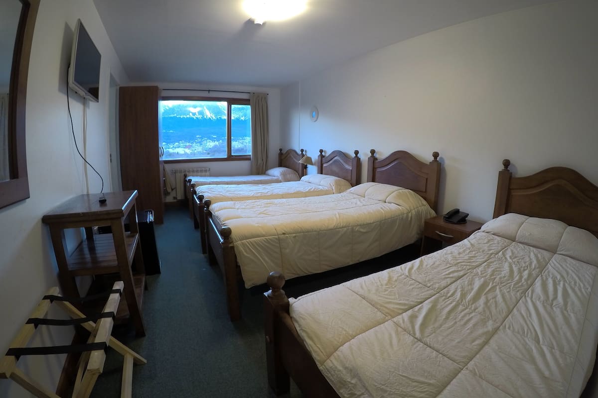 A hotel room with three single beds, a wooden desk, a TV mounted on the wall, and a window with a view of a distant landscape.
