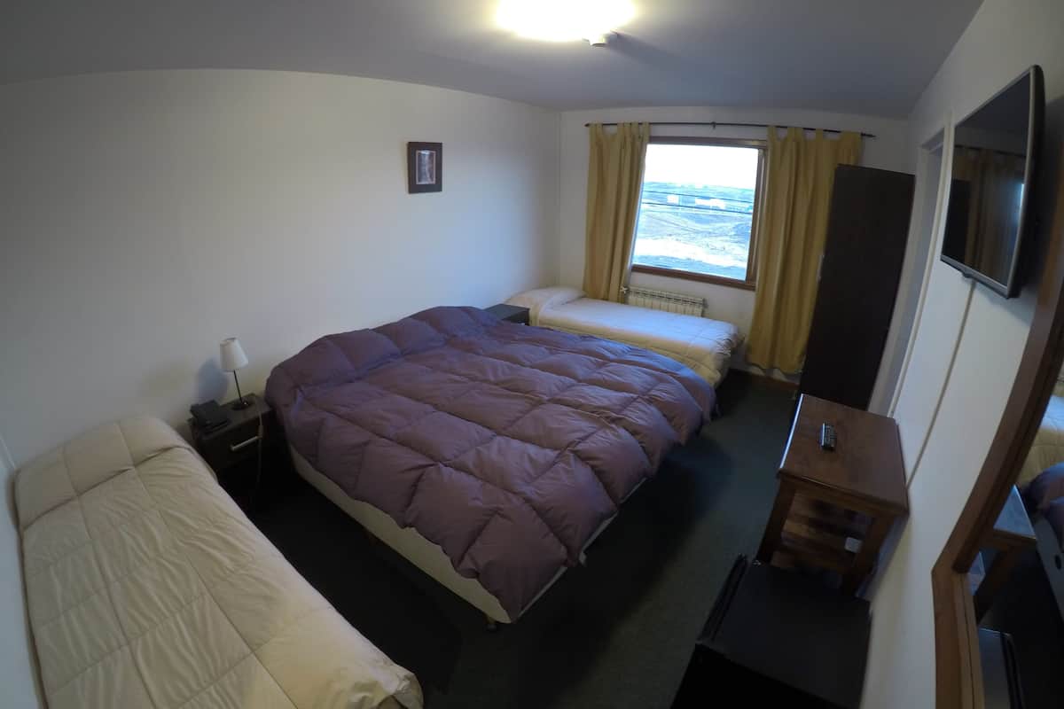 A hotel room with two beds, a bedside table with a lamp, a small table with a remote control, a window with curtains, and a mounted TV.