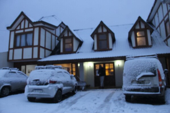A building with a traditional architectural style, featuring multiple windows and a snow-covered roof. There are several cars parked in front, also covered in snow.