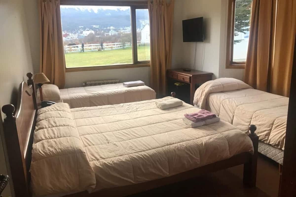 A room with two beds, a window with a view of a green landscape and mountains, a flat-screen TV on the wall, and a wooden nightstand.