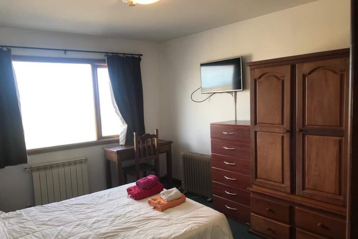 A bedroom with a bed, a wooden wardrobe, a desk, a chair, a radiator, and a TV mounted on the wall.
