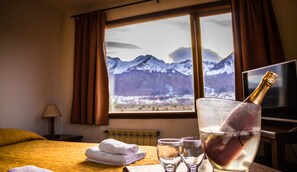 A hotel room with a bed, a window with a mountain view, a bottle of champagne, and glasses on a table.