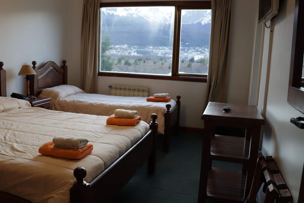 A hotel room with two beds, a small table, a chair, and a window with a view of a mountainous landscape.