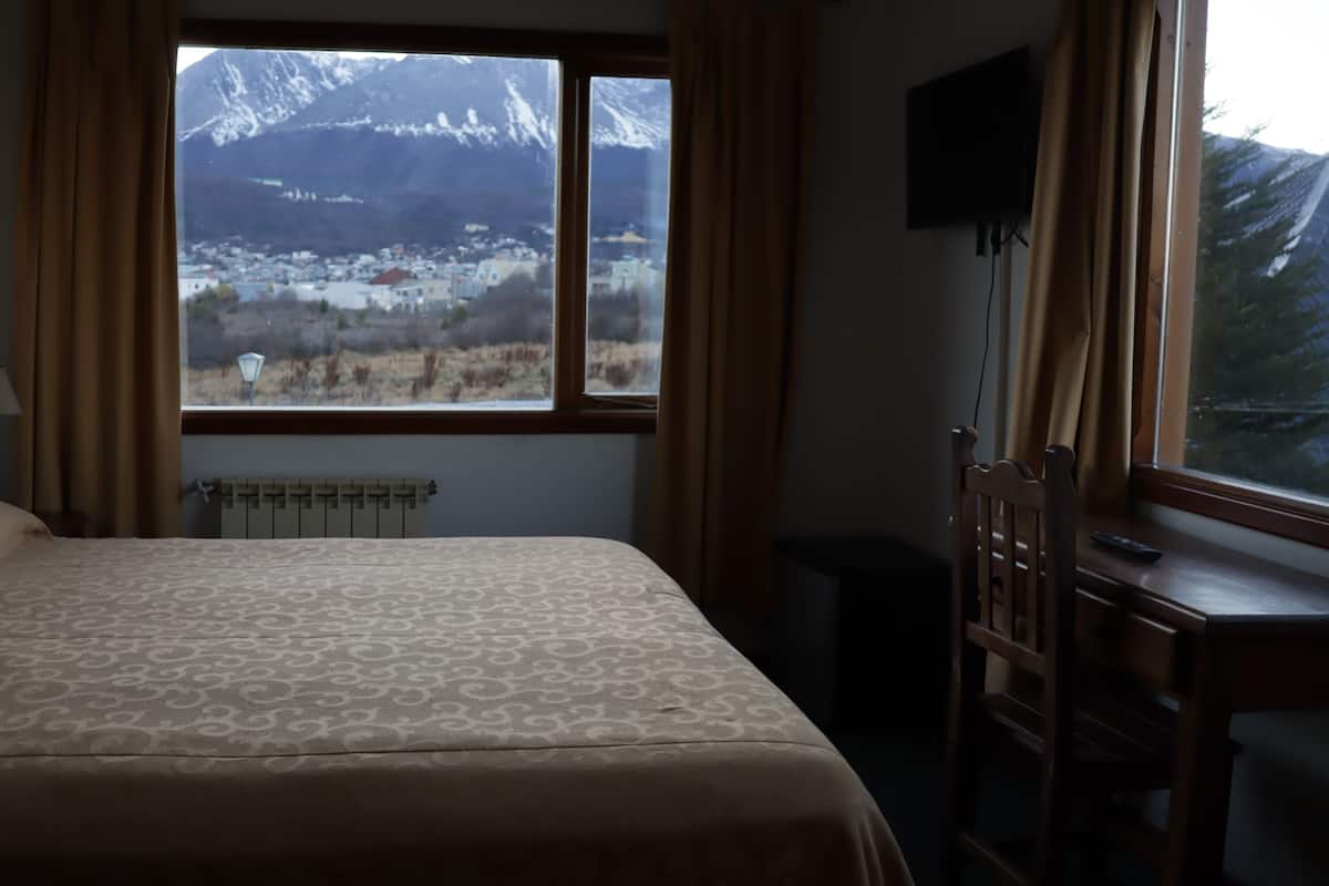 A hotel room with a bed, a desk, a chair, a radiator, and a window with a view of snow-capped mountains.