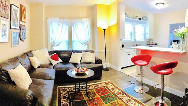 Ein Wohnzimmer mit einem dunkelbraunen Ledersofa, einem Couchtisch und einem roten Barhocker. Im Hintergrund ist eine Küche mit weißen Schränken und einer roten Barbude zu sehen.