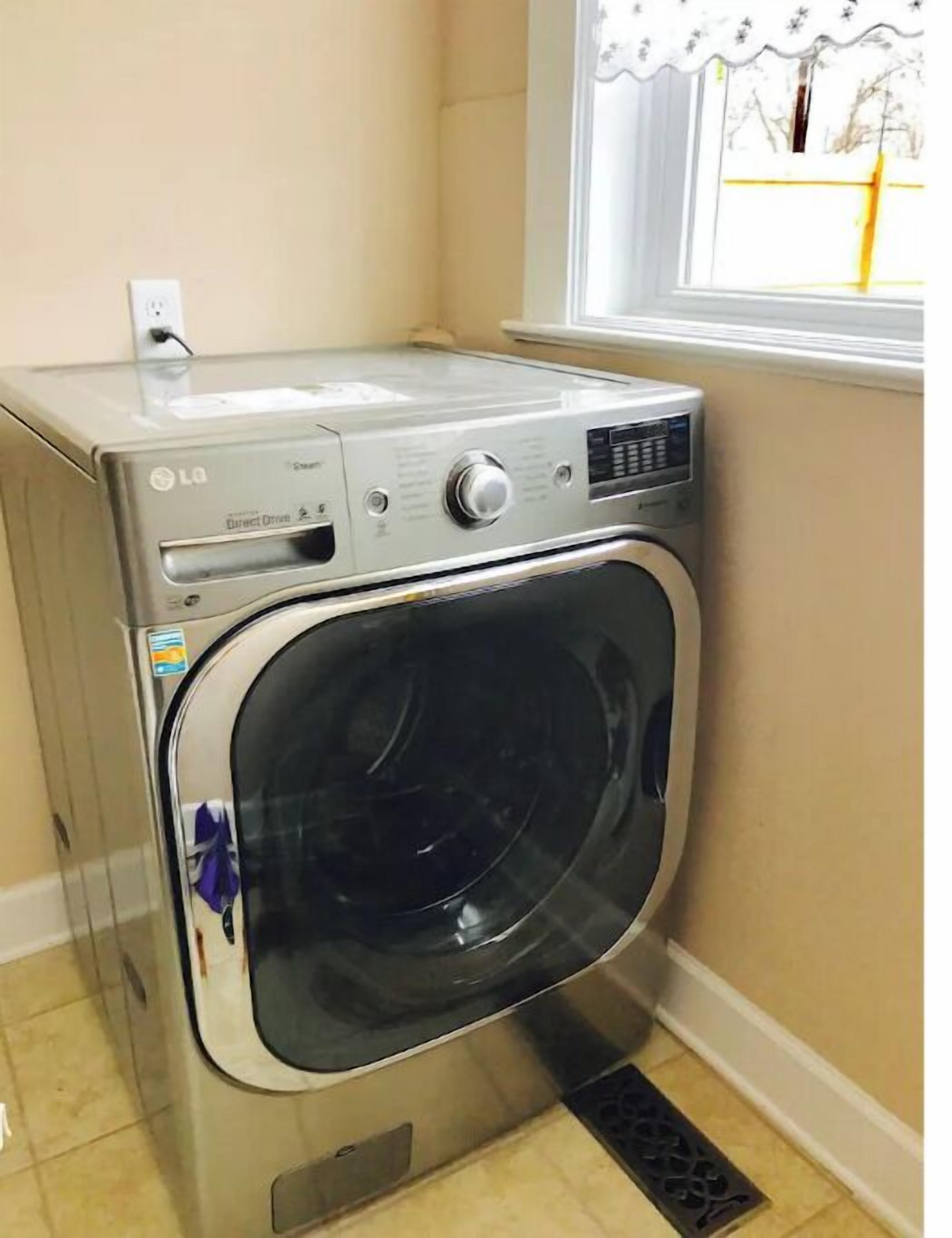 A compact, stainless steel LG Direct Drive front-loading washing machine in a corner of a room with beige walls and a window with a white frame.