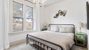 Une chambre avec une grande fenêtre, un lustre, un lit avec une tête de lit, deux tables de chevet et des éléments de décoration murale.