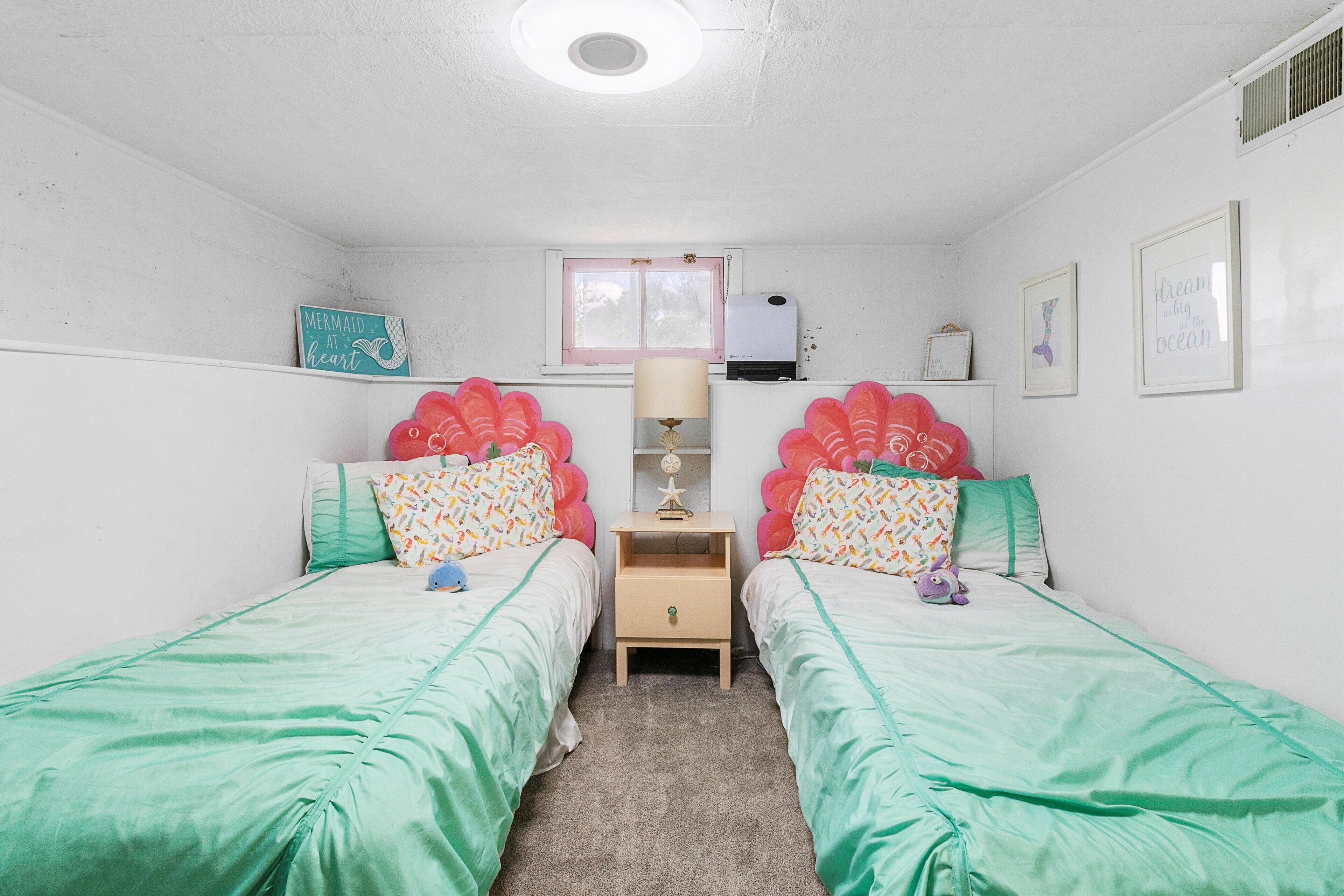 Quarto com duas camas de solteiro, mesa de cabeceira, luminária, janela e decorações nas paredes.