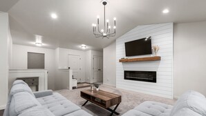 A modern living room with a large sectional sofa, a wooden coffee table, a flat-screen TV mounted on a white wall, and a fireplace below the TV.