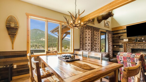 A wooden dining table with chairs, a chandelier, and a television in a room with wooden beams and a mountain view. - Show Me Mountain Views -- 20-ncd0130 3 Bedroom Home by RedAwning (Estes Park)