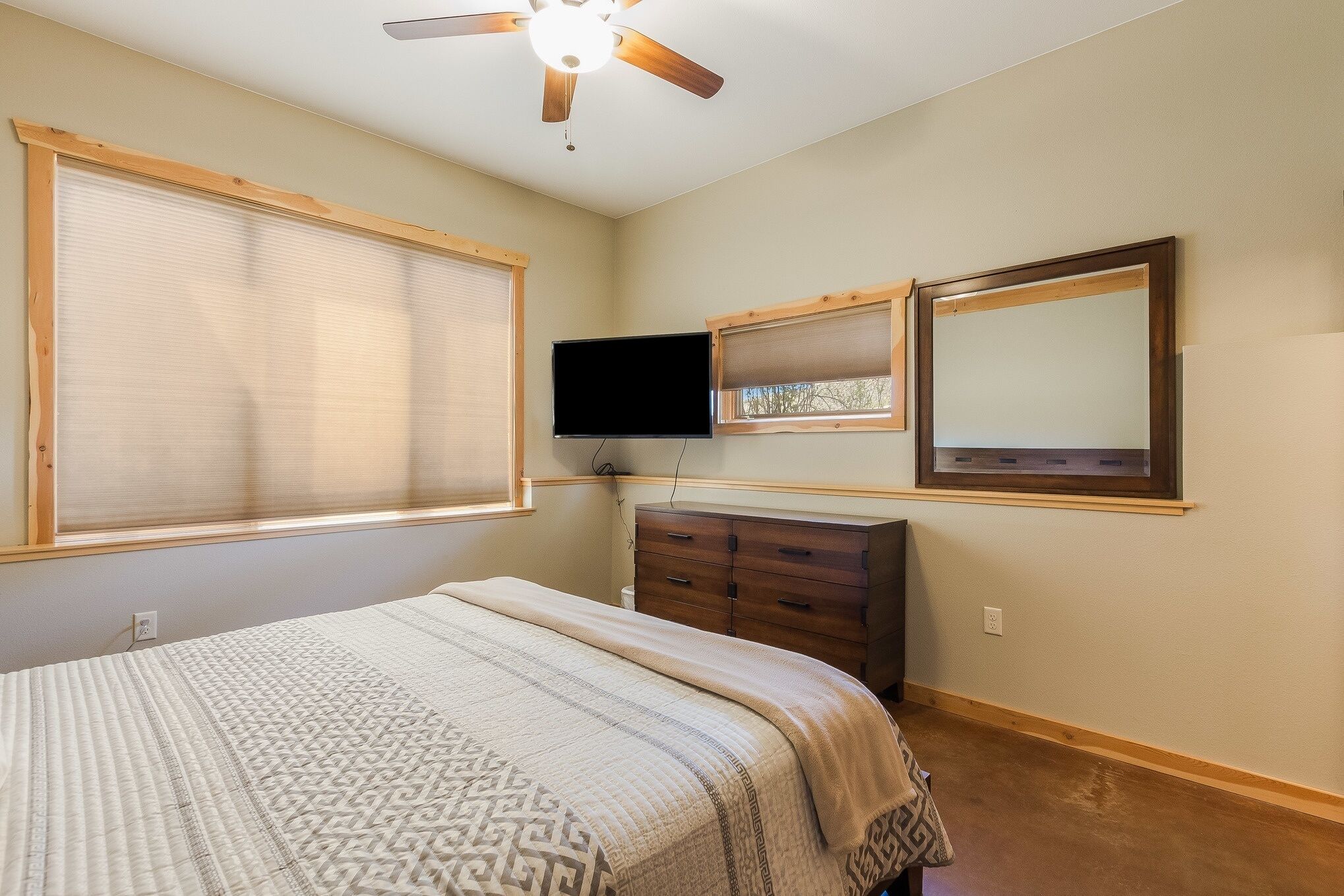 A bedroom with a bed, a dresser, a ceiling fan, a TV, and two windows with blinds.