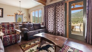A cozy living room with a leather sofa, a patterned armchair, and a wooden coffee table. A large window offers a view of a mountainous landscape. - Show Me Mountain Views -- 20-ncd0130 3 Bedroom Home by RedAwning (Estes Park)