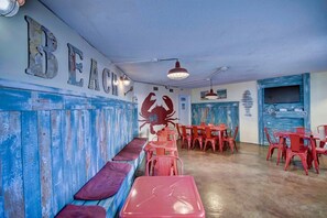 Interior de un restaurante con temática playera, que incluye un largo banco para sentarse, mesas rojas y un mural de cangrejos en la pared.