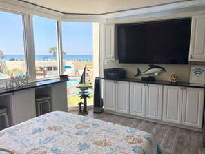 Un dormitorio con un ventanal grande, una cama con colcha estampada, un escritorio con una computadora portátil, un televisor y una decoración con delfín.