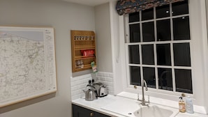 Eine Küche mit einem weißen Spülbecken, einem Fenster mit einer gemusterten Gardine, einer Karte an der Wand und einem Holzregal mit Küchenutensilien.