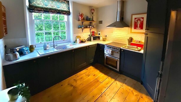 Eine Küche mit dunklen Schränken, Holzboden, einem Fenster mit Jalousien, einer Mikrowelle, einem Backofen und einem Spülbecken.