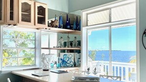 Una cocina con un ventanal grande con vistas al mar, sillas de mimbre blancas y armarios de madera.