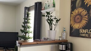 Un ambiente compatto con un tavolo di legno, una macchina da caffè, una piccola pianta e uno scaffale con bottiglie e una sveglia appesa al muro.
