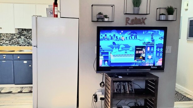 A living room with a flat-screen TV mounted on the wall, a wooden entertainment center below, and a kitchen area with cabinets and a refrigerator in the background.