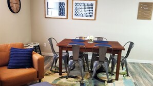 A dining area with a wooden table, metal chairs, a blue cushion on a leather sofa, and abstract art on the wall.