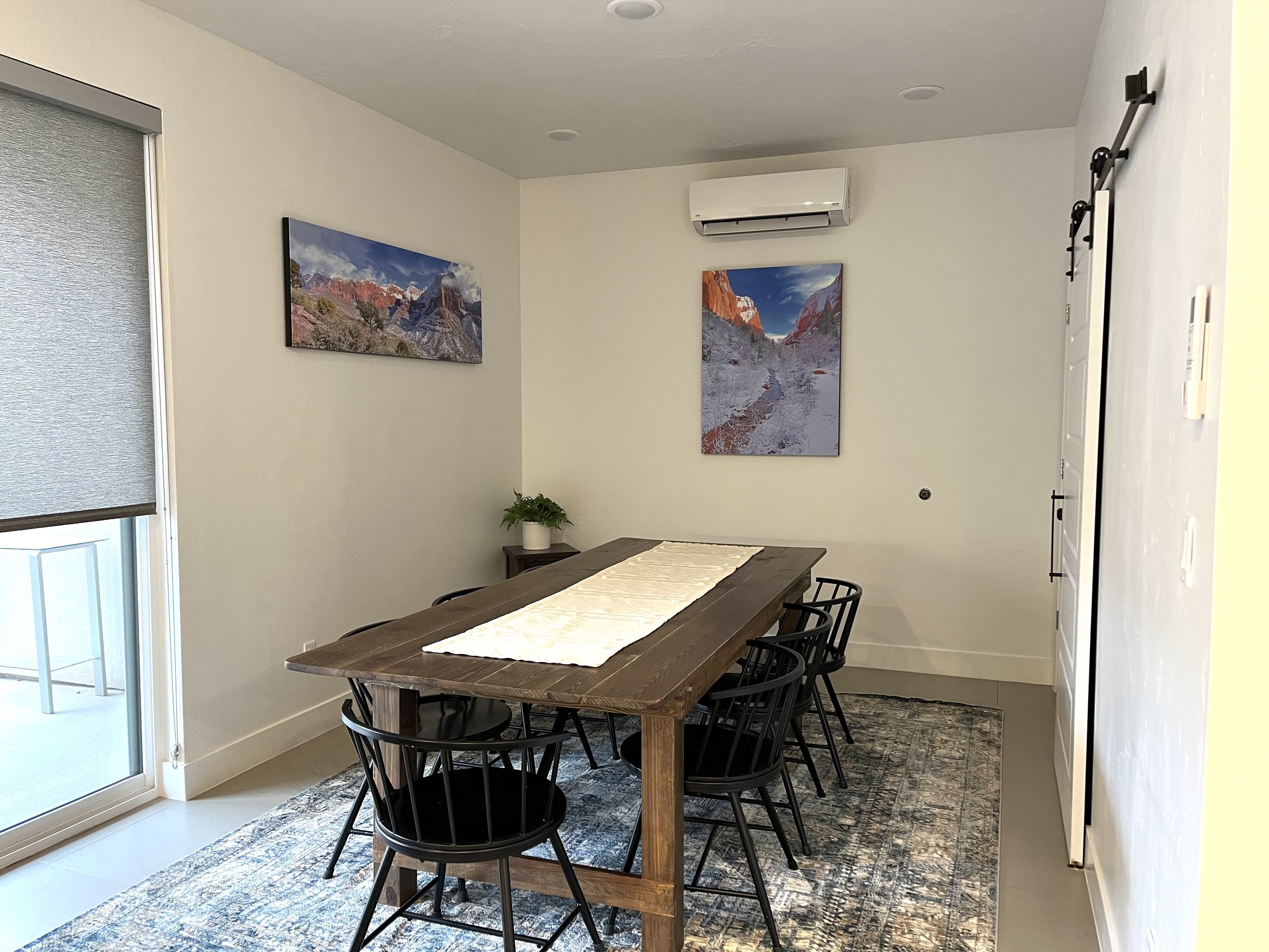 Een vergaderruimte met een lange tafel, zwarte stoelen, twee schilderijen, een airconditioner en een plant in pot.