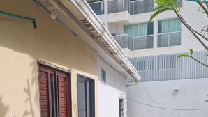 Een gebouw met een witte gevel, een rode deur en een klein balkon. Planten in de voorgrond en een betonnen pad dat naar het gebouw leidt.