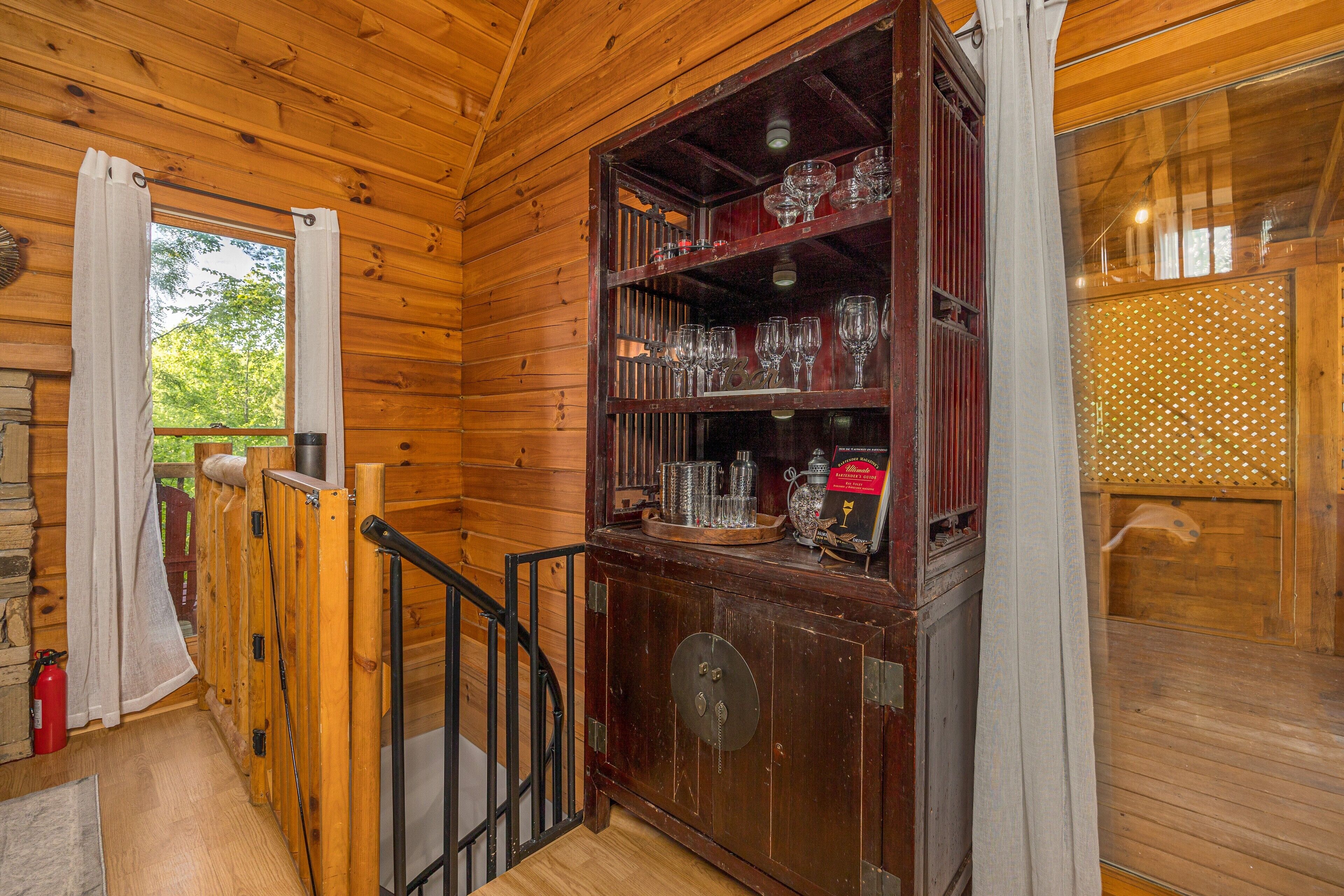 Interior de una cabaña de madera con una escalera de madera, un ventanal que da al exterior y un mueble de madera con cristalería.