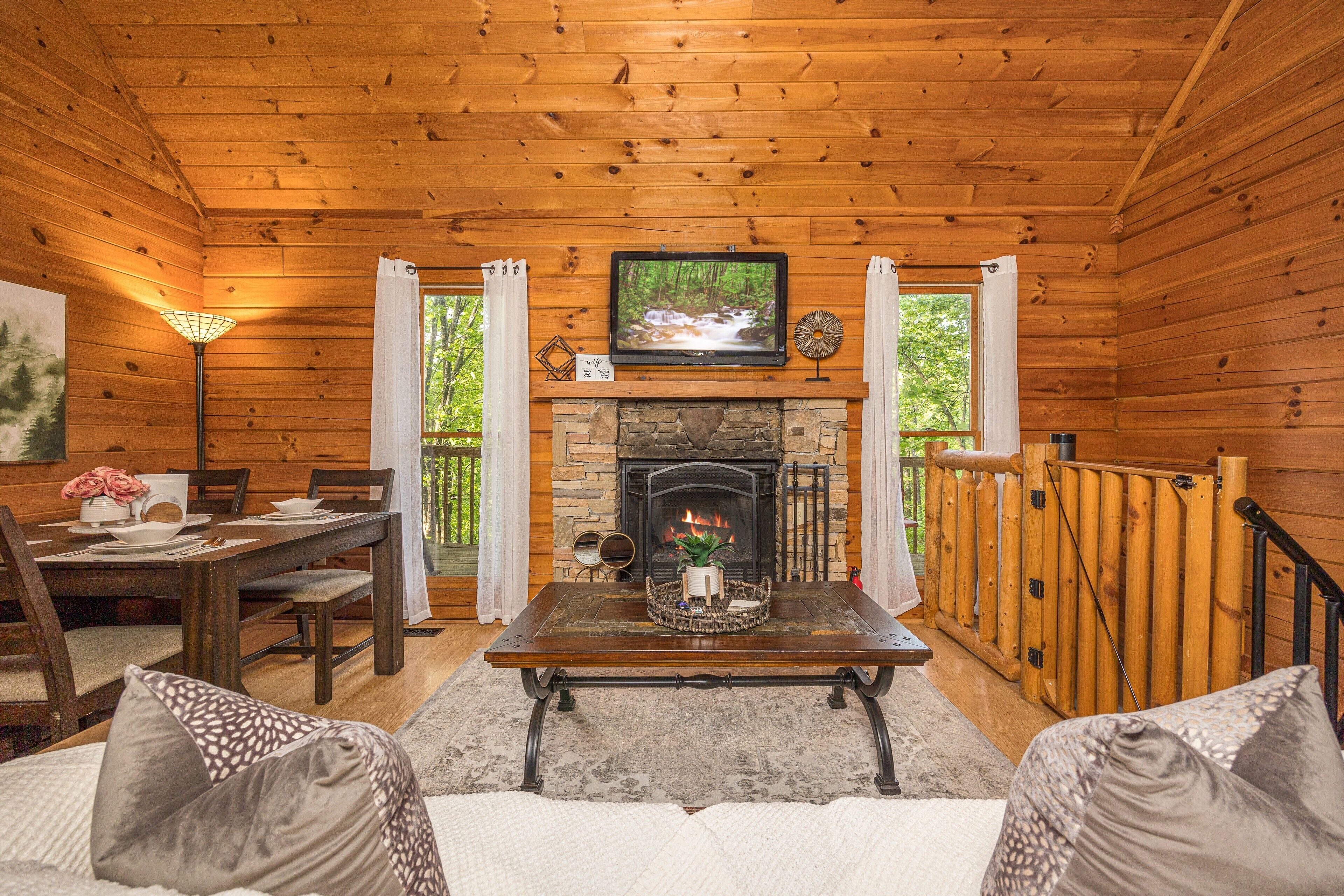 A cozy cabin interior with a stone fireplace, wooden walls, a dining area, and a TV mounted above the fireplace.