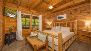 A wooden cabin bedroom with a bed, a television, a fireplace, and a window with curtains.