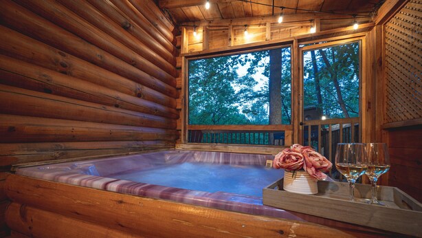A wooden cabin interior with a hot tub, a window with a view of trees, and a tray with a vase and glasses.